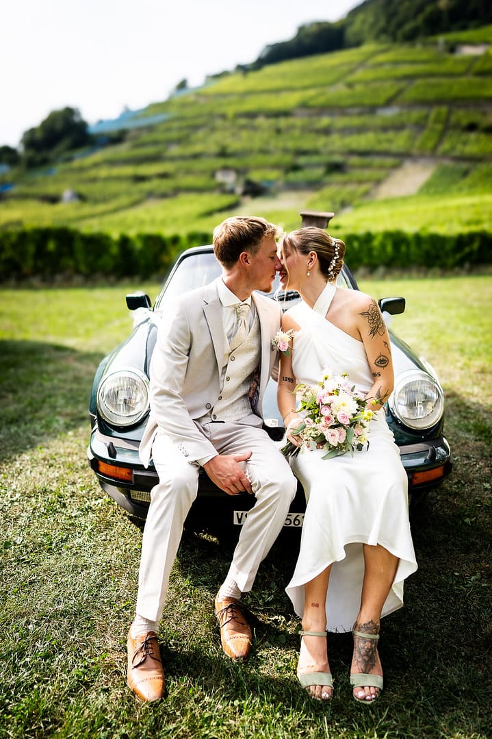 Mariage dans les vignes : La dolce vita d'Aurélien & Claudia Au cœur des vignobles vaudois, Aurélien et Claudia s'offrent une parenthèse rétro et romantique. Assis sur leur Alfa Romeo, entourés par la poésie des coteaux, ils échangent un baiser complice face à l'immensité du paysage. Un instant de complicité vintage capturé par Dis-Moi Oui, où l'élégance italienne rencontre le charme authentique du terroir vaudois.
